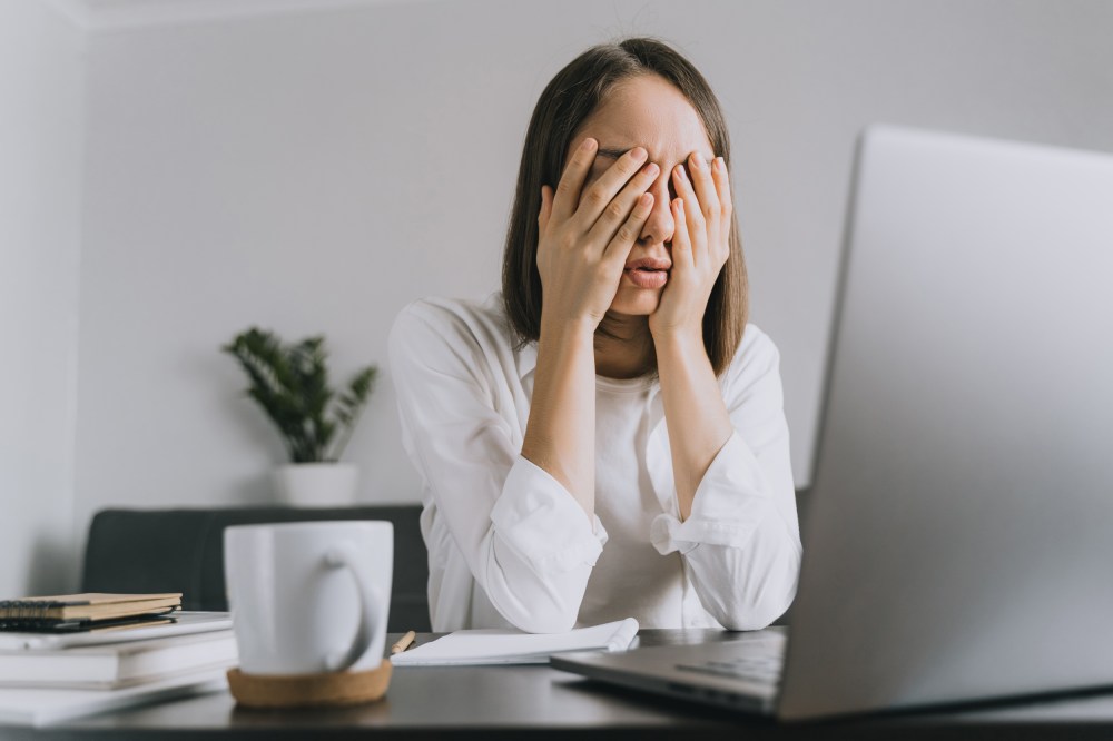 A woman sitting at desk holds her head in her hands.