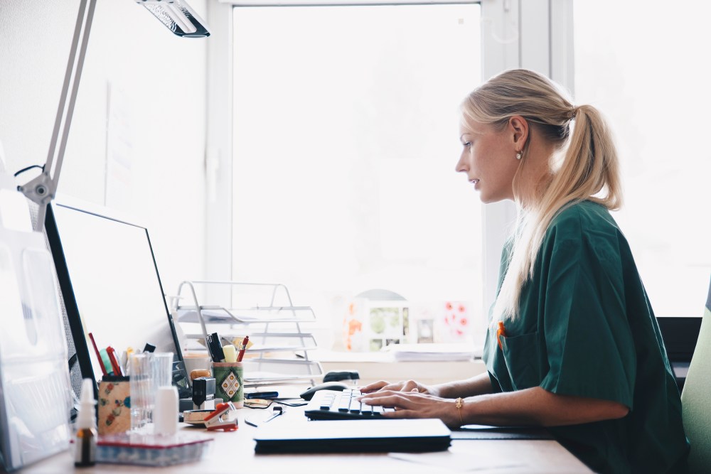 Image: Side view of female nurse working at computer desk against window in office