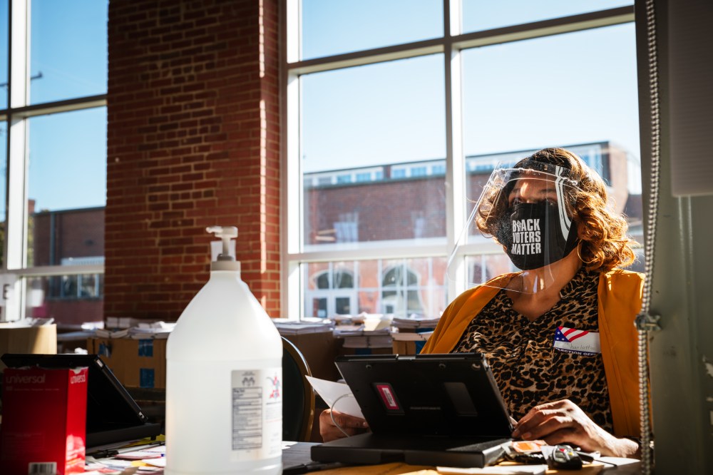 Image: An election official wearing a mask that reads "Black Lives Matter" sits at her desk speaking to a voter