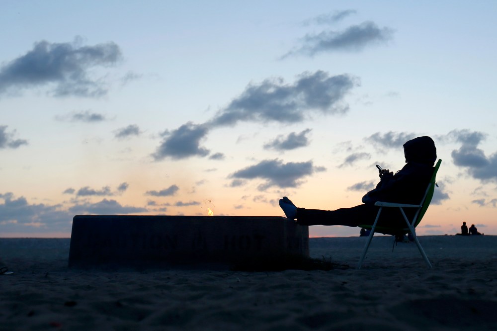 A woman sits by an open fire at Moonlight Beach as she reads messages on her phone after sunset in Encinitas, California, March 31, 2014.