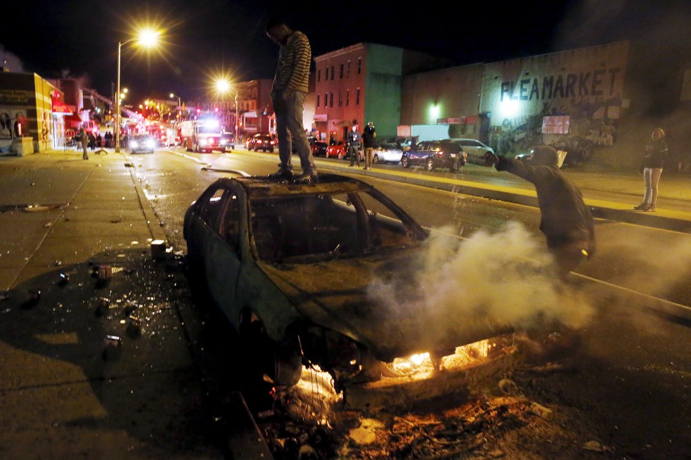 A rioter stands atop a burning car as another man pours fuel onto the fire while Baltimore firefighters behind them fight fires in mutliple burning buildings set ablaze by rioters during clashes in Baltimore, Md., April 27, 2015. (Jim Bourg/Reuters)