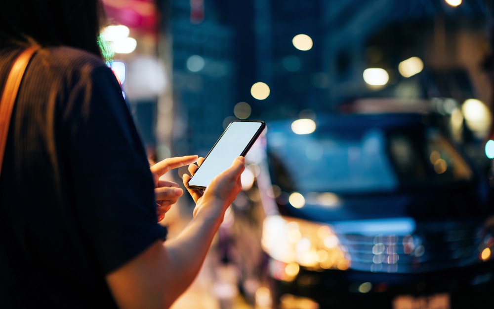 Close up of young woman using mobile app device on smartphone to hail a taxi ride on city street after work in the evening.