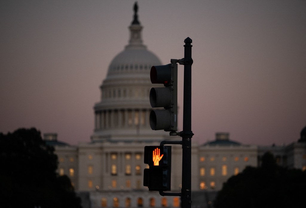 A crosswalk signal of a traffic light flashes an orange hand backdropped by the Capitol.