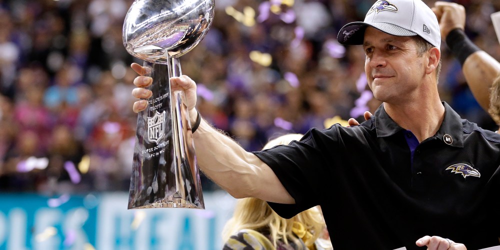 John Harbaugh holds the Vince Lombardi Championship trophy as purple and gold confetti falls in the background.