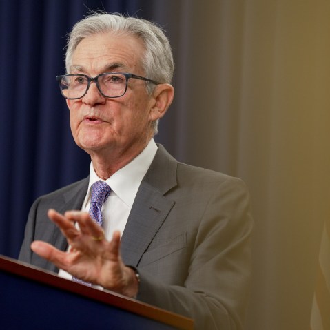 Jerome Powell lifts his left hand while speaking at a podium during a news conference. The photo is shot from below.