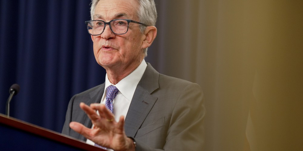 Jerome Powell lifts his left hand while speaking at a podium during a news conference. The photo is shot from below.