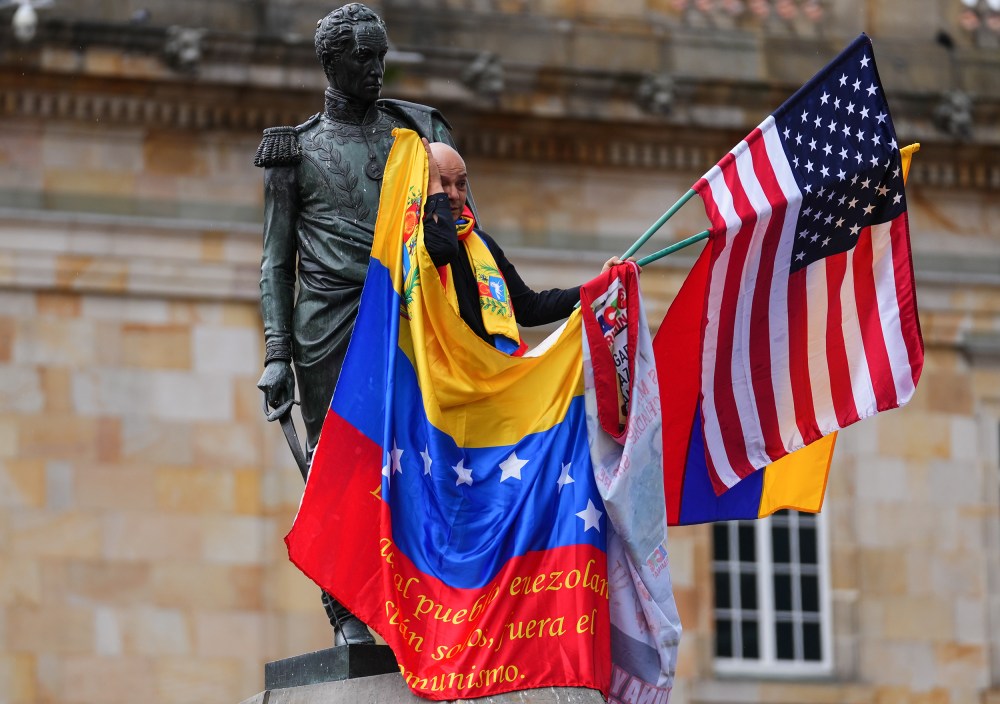 A man waves a venezuelan and american flag while standing a statue in Bogota.