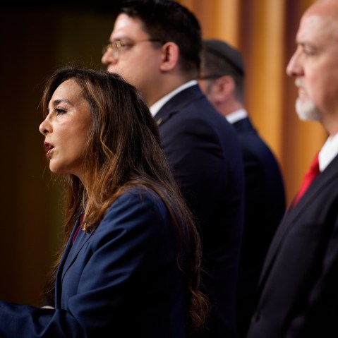 Harmeet Dhillon speaks at a podium. This is a profile shot. She is surrounded by aides.