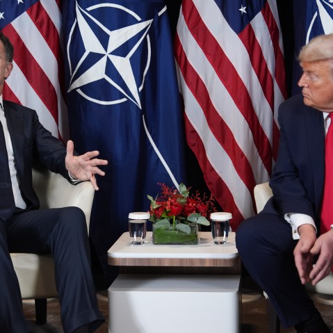 Mark Rutte (left) and Donald Trump sit next to each other in front of flags for the U.S. and NATO. Trump looks at Rutte as he speaks.