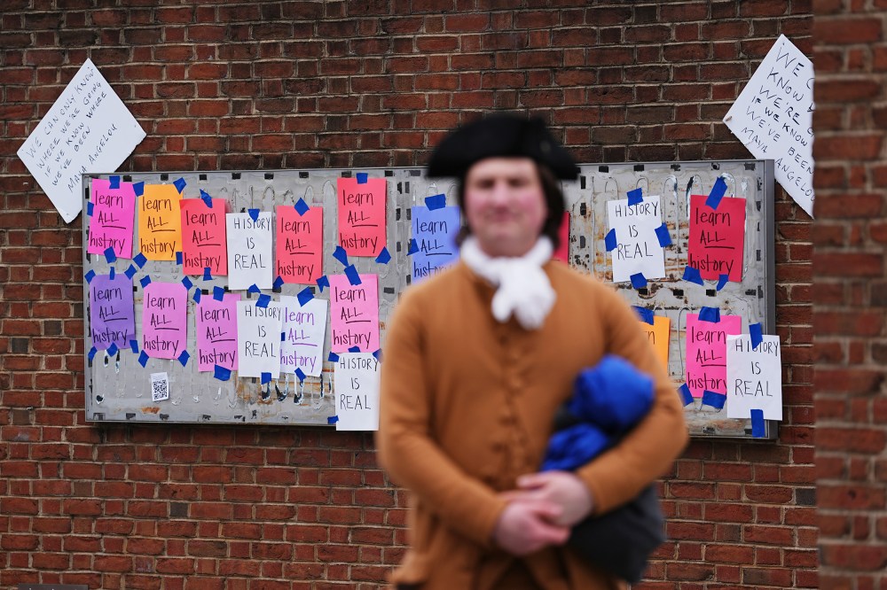 A person dressed in a late 18th- or early 19th- century costume stands blurred in the foreground. In focus in the background, multicolored paper signs that read "learn ALL history" and "HISTORY IS REAL" are taped to an empty panel on a brick wall.