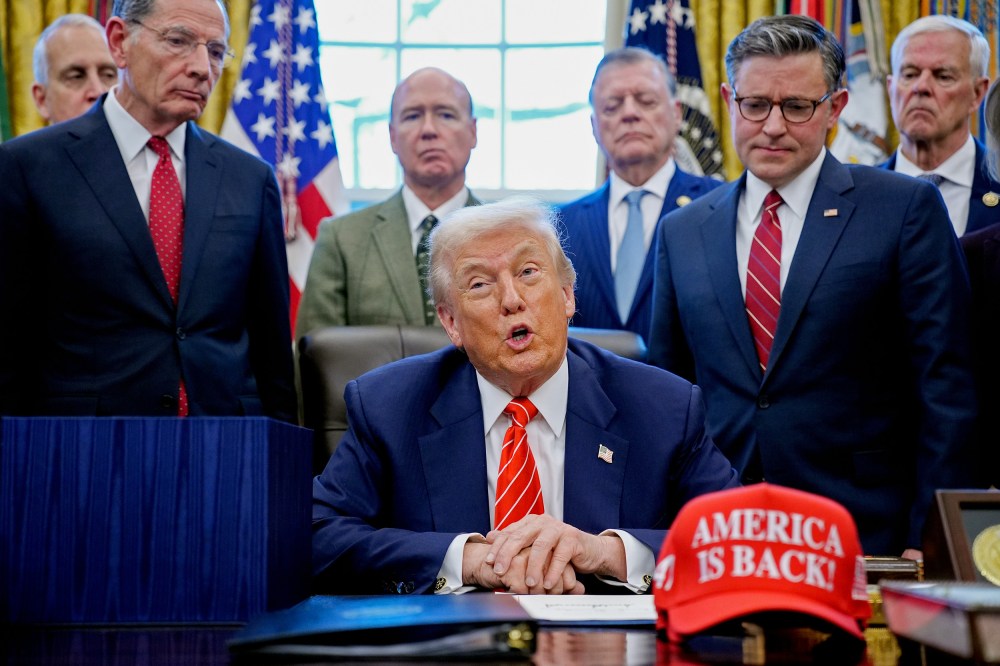 President Donald Trump, center, surrounded by members of the House and Senate in the Oval Office. Out of focus on the desk is a red hat that says "AMERICA IS BACK!"