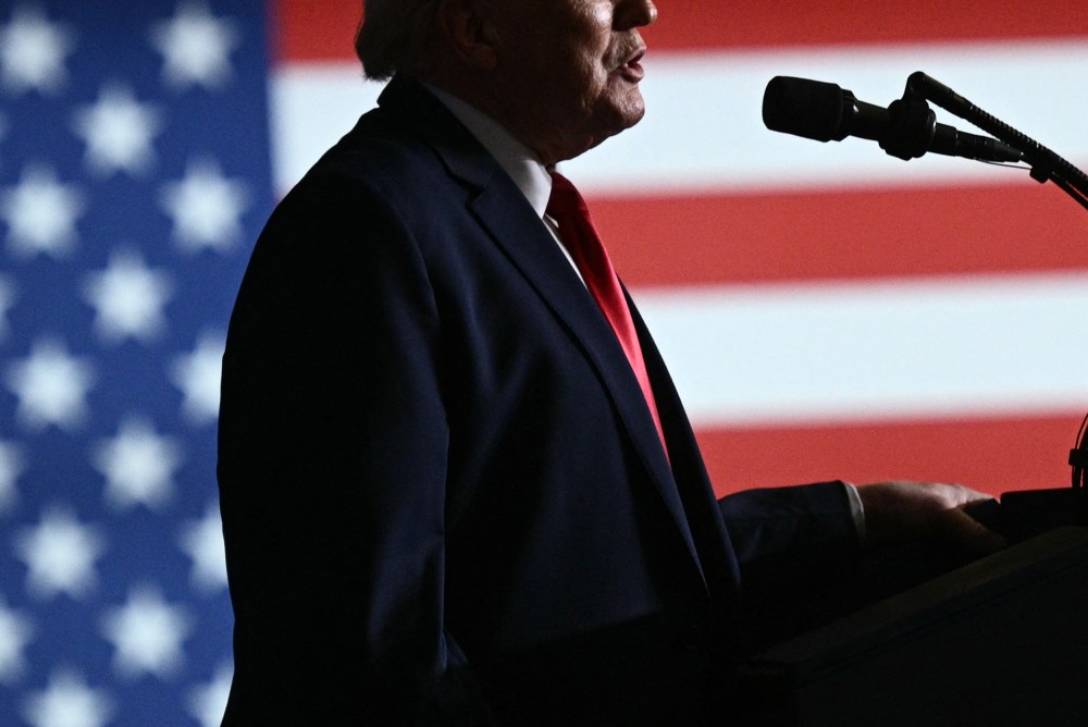 Donald Trump's mouth and torso in profile speaking into a microphone. Behind him, the American flag.