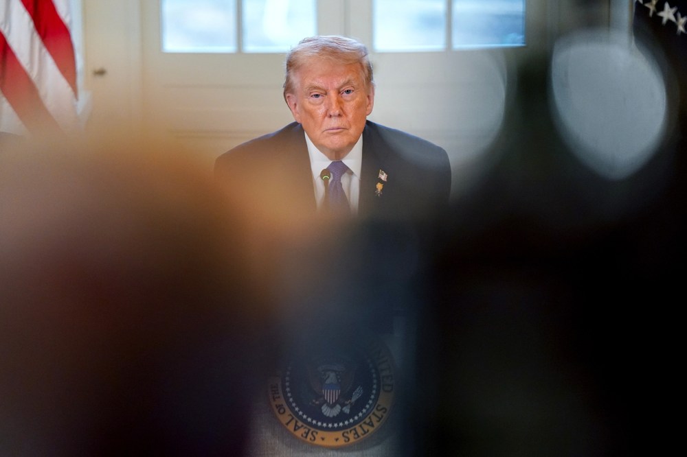 Donald Trump looks on, partially obscured by people in the foreground.