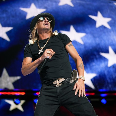 Kid Rock holds a microphone on stage. The background is an American flag.
