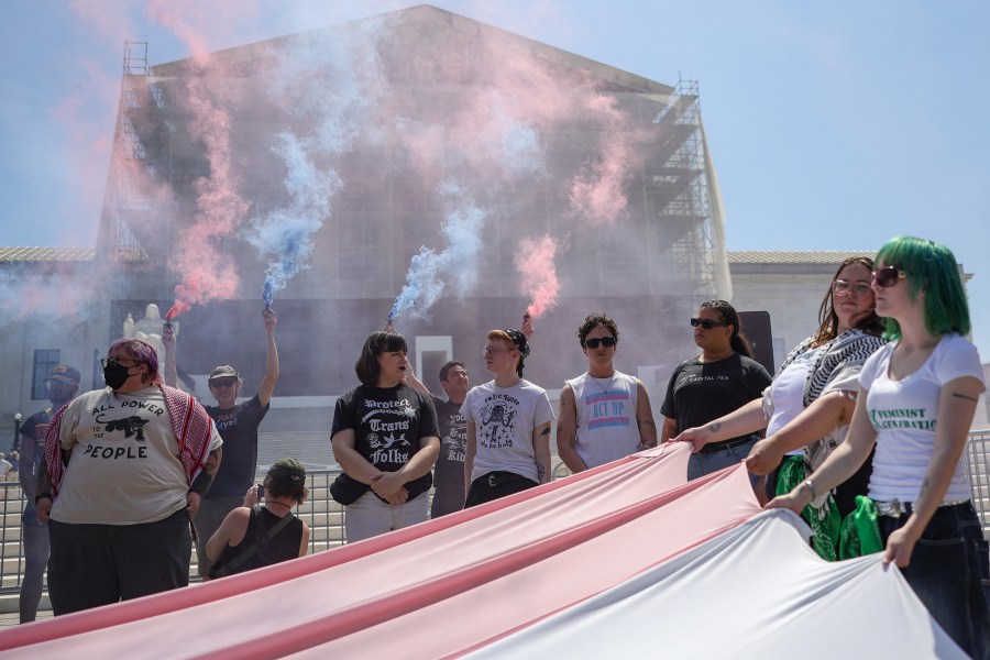 People hold pink and blue colored smoke devices in front of the Supreme Court. People in the foreground hold corners of large pink and blue banners parallel to the ground.