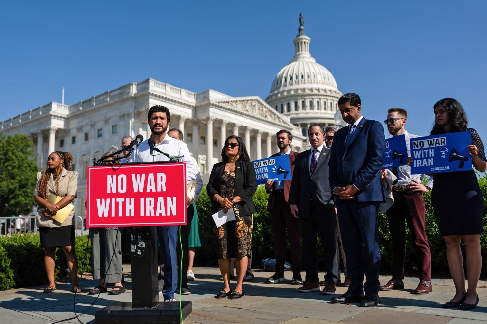 Greg Casar stands behind a podium that has a red sign that says "NO WAR WITH IRAN." He is surrounded by other people, and in the background is the Capitol.