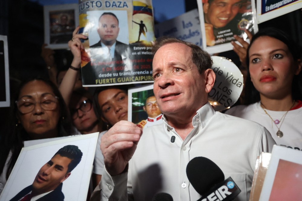 Juan Pablo Guanipa stands in front of a microphone with a crowd of people behind him holding signs for the release of other Venezuelan prisoners.