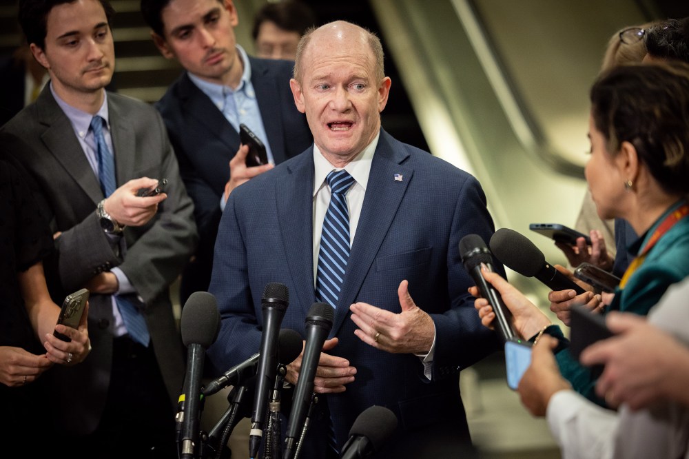 Chris Coons speaks while surrounded by reporters holding phones and microphones.