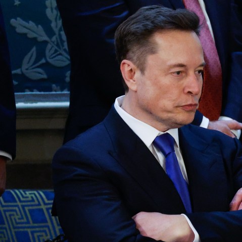 Elon Musk looks down and has his arm crossed on his chest.