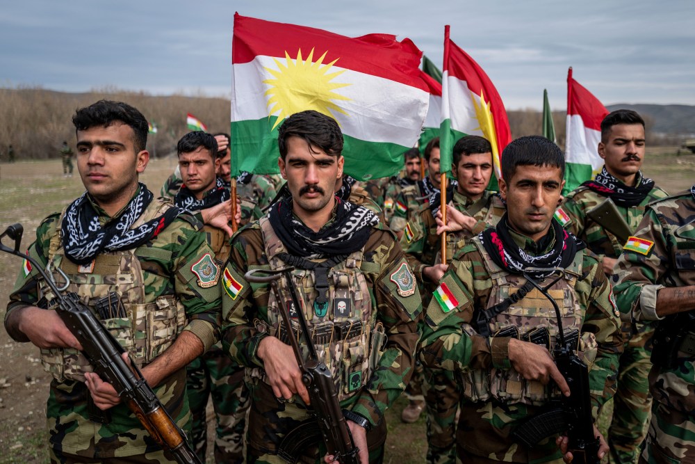 People in military uniforms holding guns and Kurdish flags stand in formation.