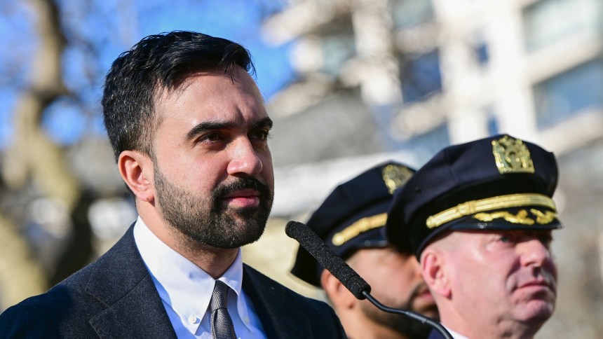 Zohran Mamdani stands in front of a microphone. Police officers are seen behind him.