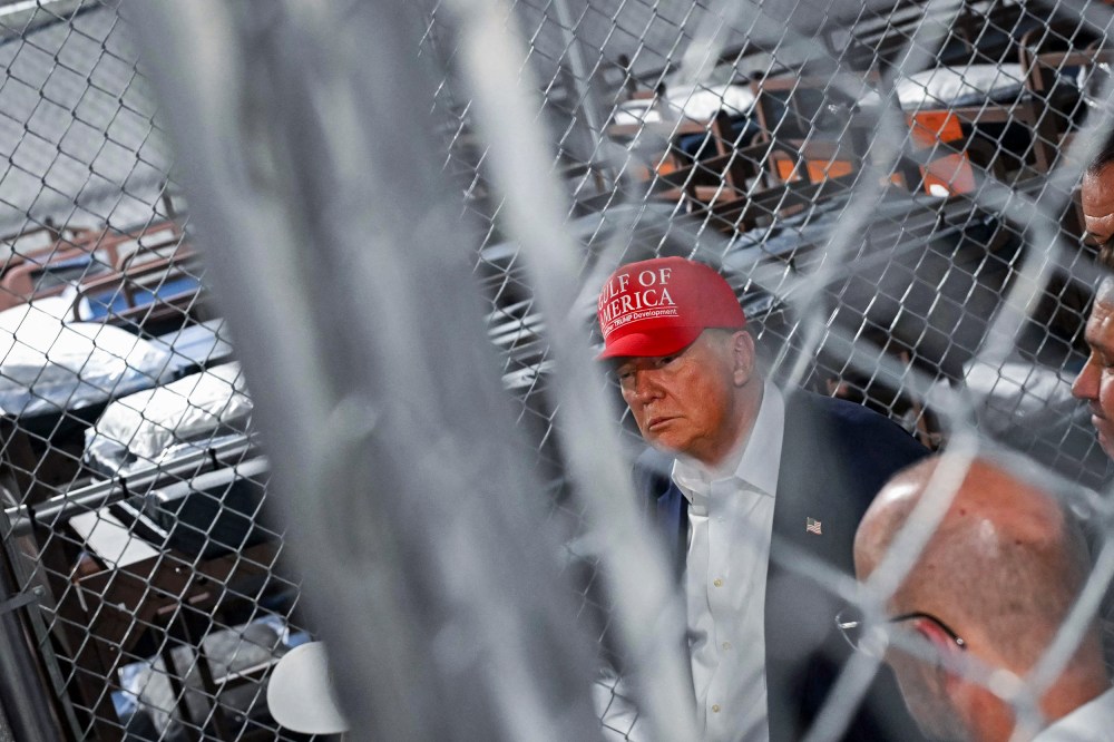 Donald Trump, wearing a red "Gulf of America" hat, is seen from behind a chainlink fence. Rows of bunk beds are seen in the background.