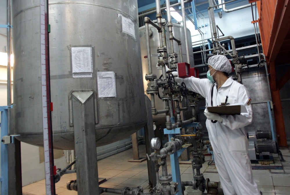 A person in a white coverall wearing a paper mask and a hairnet has an arm out toward big machinery while the other arm holds a clipboard.