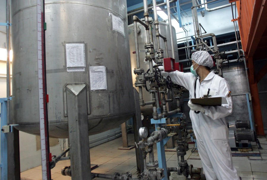 A person in a white coverall wearing a paper mask and a hairnet has an arm out toward big machinery while the other arm holds a clipboard.