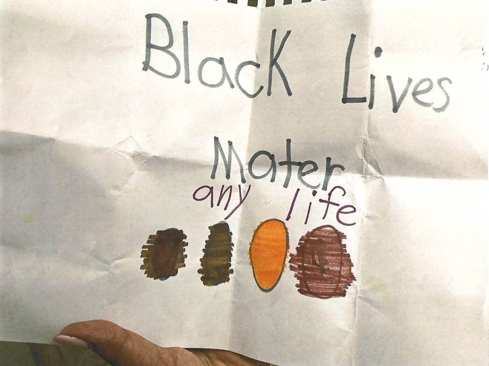 A person's hand is seen holding an unfolded piece of paper. The paper, drawn on with marker, says, "Black Lives Mater any life." Below it are four ovals colored in with different shades of brown.