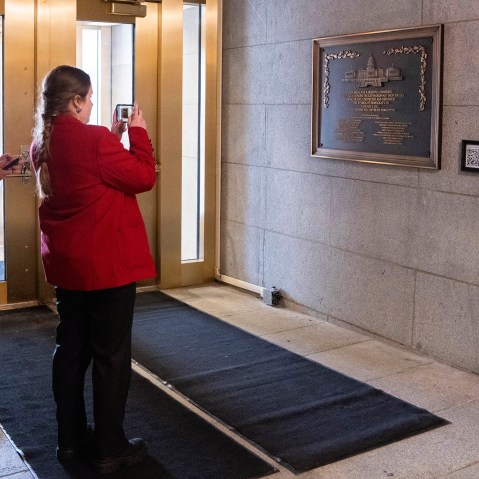 Two tour guides wearing black pants and red jackets take photos of the plaque.