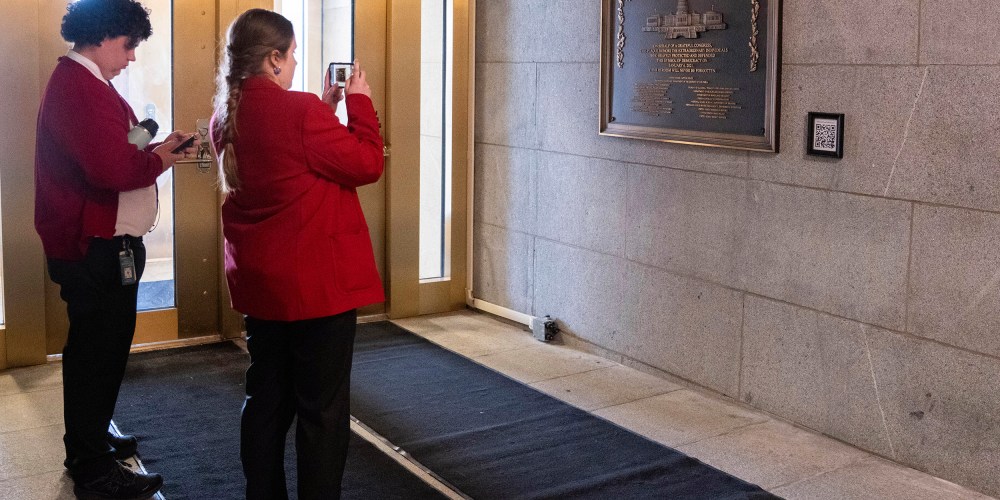 Two tour guides wearing black pants and red jackets take photos of the plaque.