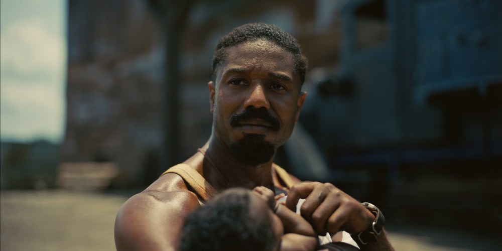 Michael B. Jordan holds a Black baby out in a space that looks like a town square.