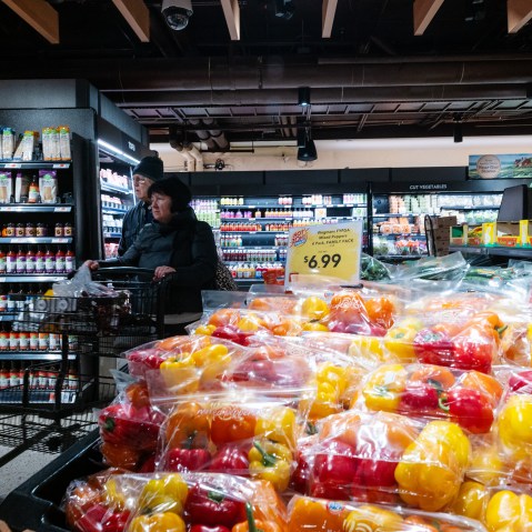 Red and yellow peppers are seen in the foreground while a woman pushes a cart in a grocery store.