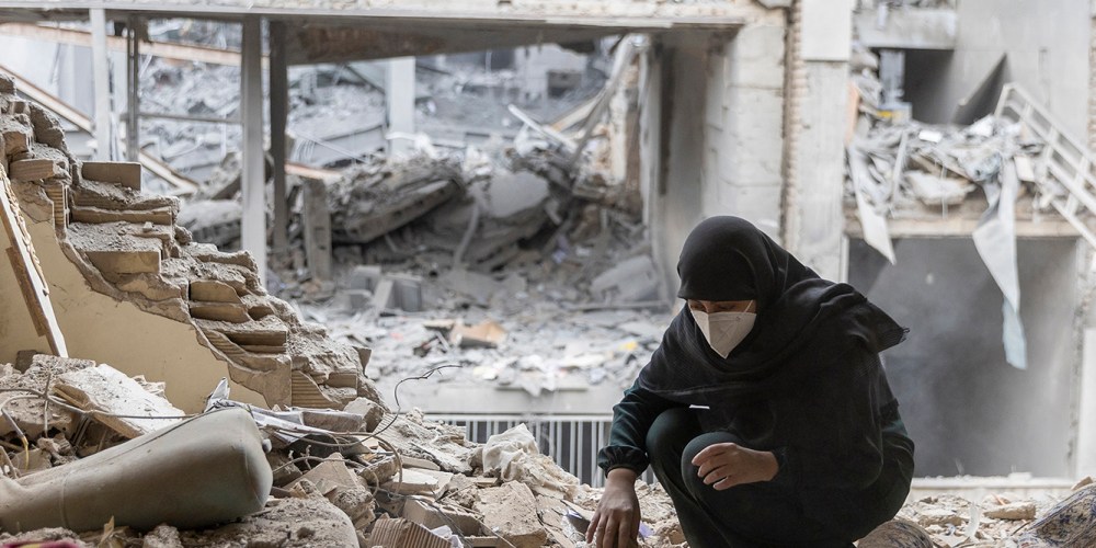 One woman wearing a black hijab sitting on the floor as she searches through the reubbles of her home. In the background, there are buildings destroyed with US-Israel attacks.