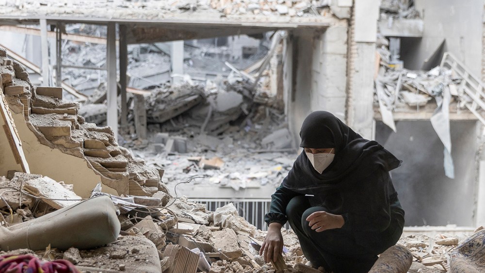 One woman wearing a black hijab sitting on the floor as she searches through the reubbles of her home. In the background, there are buildings destroyed with US-Israel attacks.