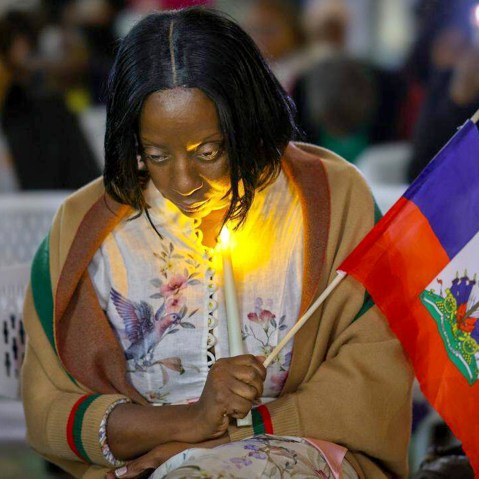 Haitian community activist bows her head down as she prays and holds a candle and Haiti flag.