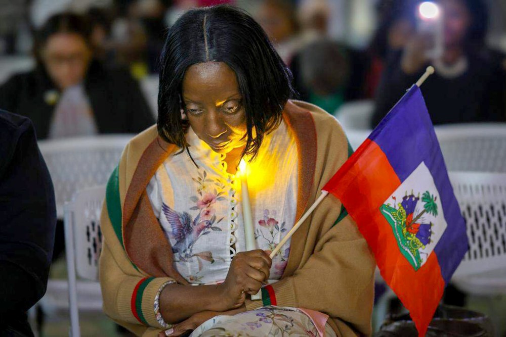 Haitian community activist bows her head down as she prays and holds a candle and Haiti flag.
