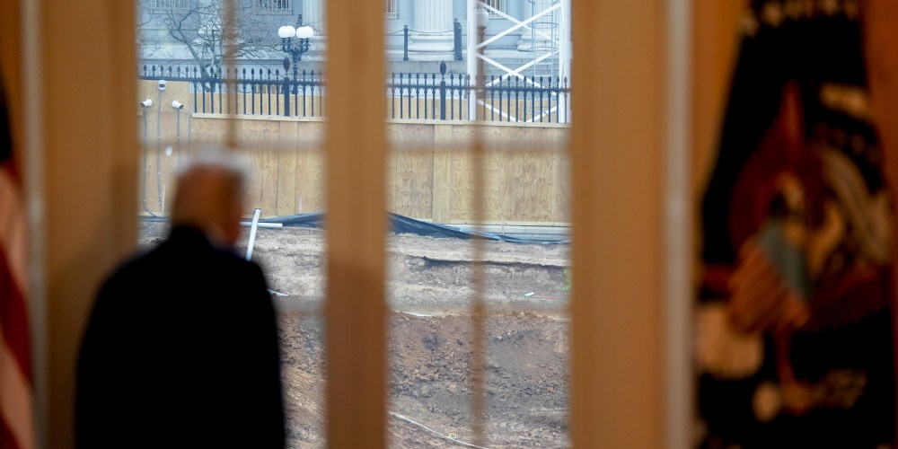 Donald Trump, out of focus in the foreground, looks away from the camera and out a set of windows, where construction is seen in focus.