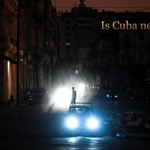 A man walks while cars cruise along a street during a blackout in Havana, Cuba. Text on top right reads “Is Cuba Next?”