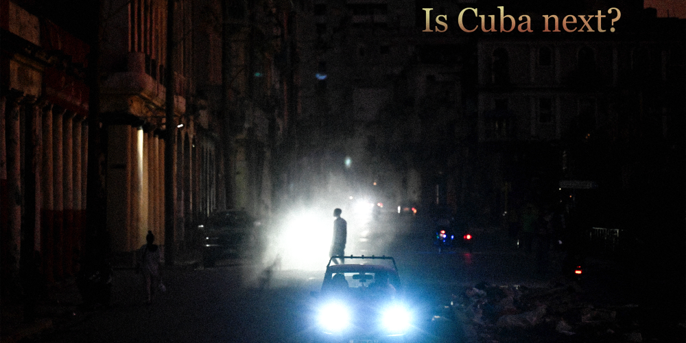 A man walks while cars cruise along a street during a blackout in Havana, Cuba. Text on top right reads “Is Cuba Next?”