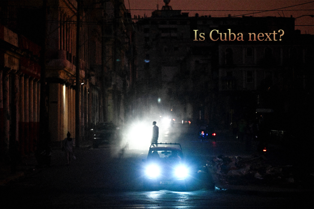 A man walks while cars cruise along a street during a blackout in Havana, Cuba. Text on top right reads “Is Cuba Next?”